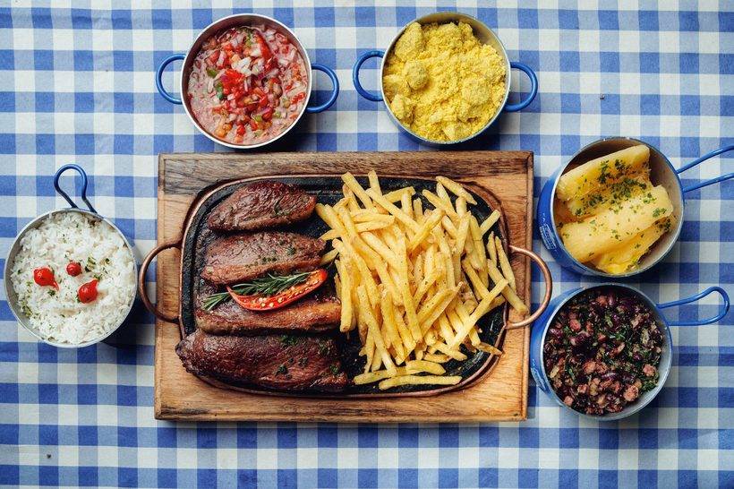 Tábua de carne com batata frita e as panelinhas de arroz, vinagrete, farofa, mandioca e feijão tropeiro em volta, sobre a toalha xadrez azul